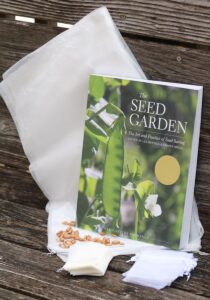 A book, large mesh lettuce bags, small mesh blossom bags, small white glassine envelops, and some brown seeds arranged on a wooden bench